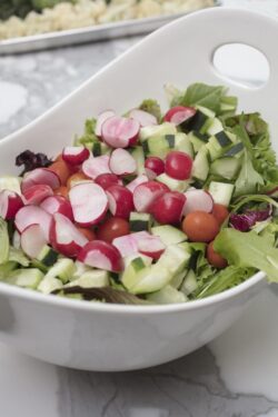 Preparation of a Salad made with greens, lettuce, radishes, cucumbers, olives, tomatoes in a white ceramic bowl.