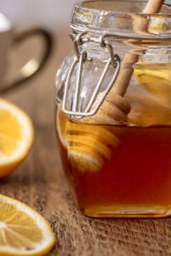 Jar of honey being dripped from a honey stick with a side of lemon on a wood table top