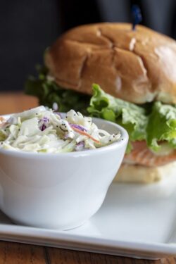 Grilled salmon on a toasted bun with lettuce and cheese, served with a small bowl of coleslaw