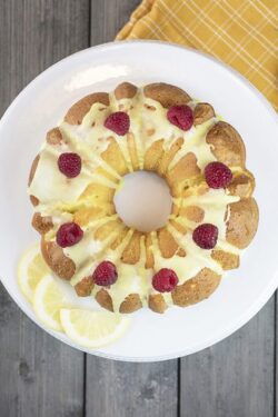Lemon cake frosted with yellow sugar icing and red raspberries, dressed with lemon slices on the side on a white platter