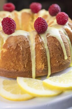 Lemon cake frosted with yellow sugar icing and red raspberries, dressed with lemon slices on the side on a white platter