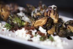 An appetizer plate of roasted Brussels Sprouts garnished with Feta cheese and bacon, with a drizzle of Balsamic Glaze