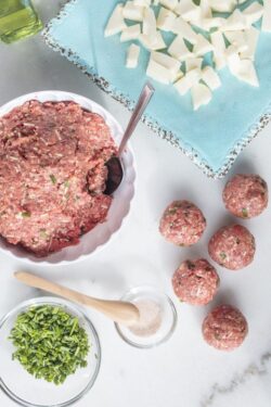 Marble table top with ingredients to make meatballs including raw ground beef, mozzarella cheese, chives and salt