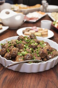Dish of meatball appetizers, mixed with garlic and cheese, with various other appetizers in the background